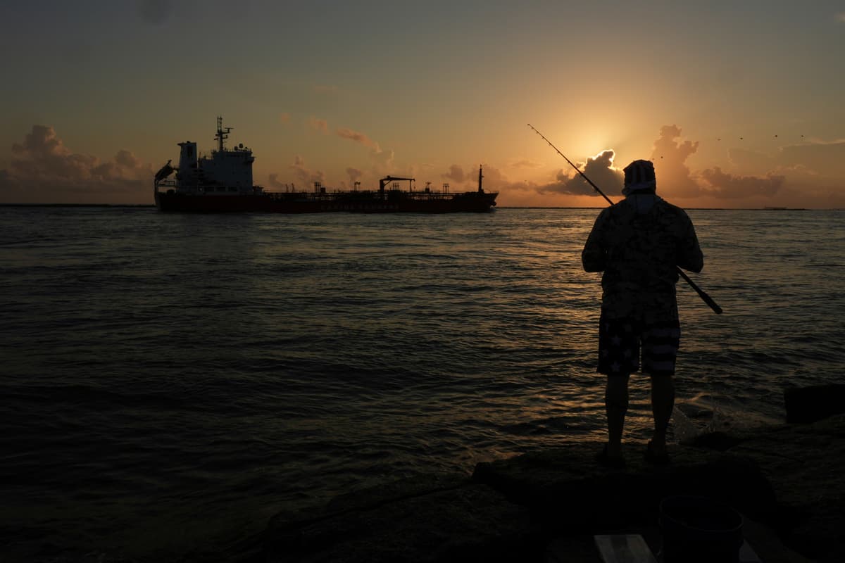 An oil tanker passes at sunrise while a man fishes in Port Aransas, Texas.