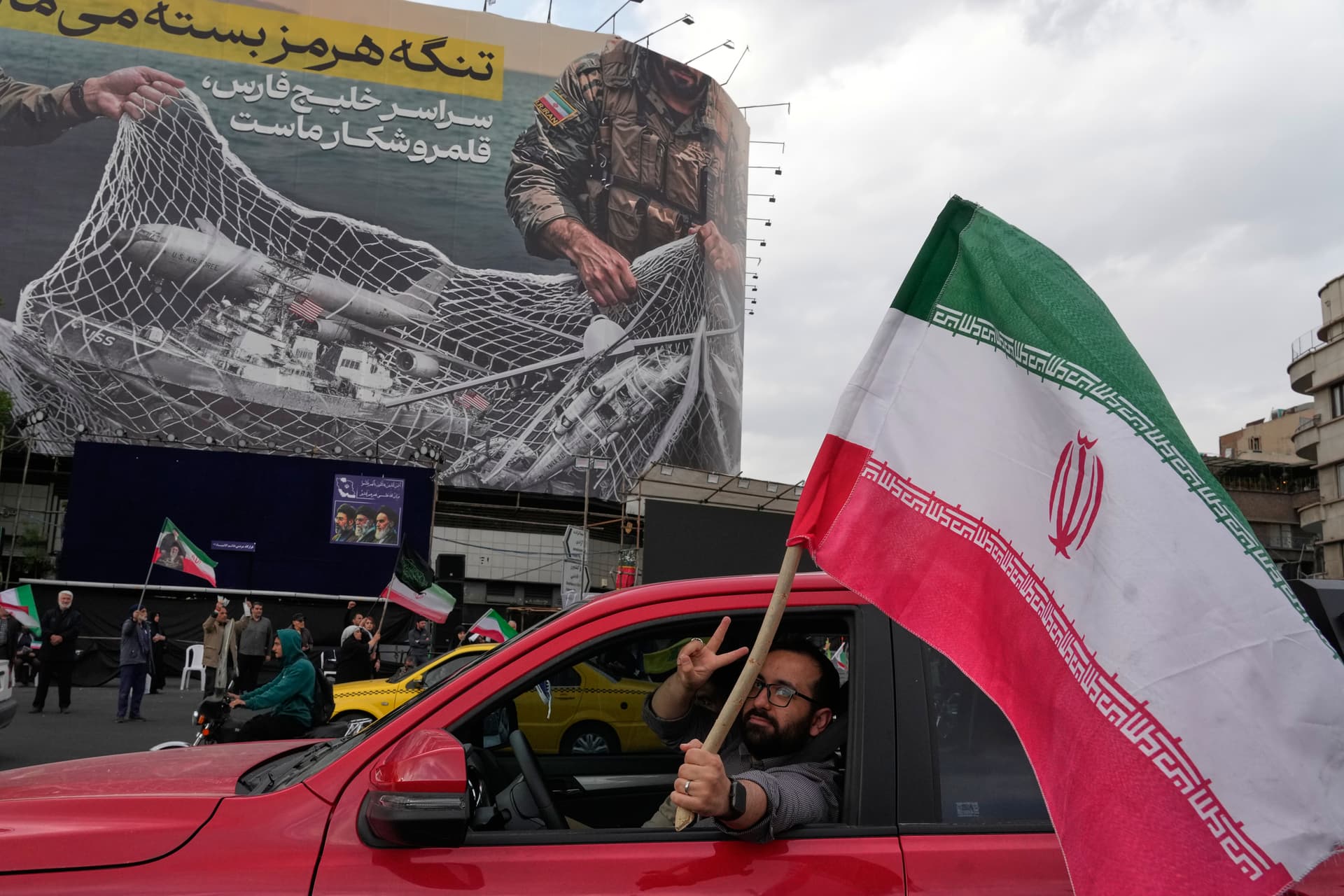 A man flashes a victory sign as he carries an Iranian flag in front of an anti-U.S. billboard that reads in Farsi: 'The Strait of Hormuz will remain closed, The entire Persian Gulf is our hunting ground,' April 13, 2026.