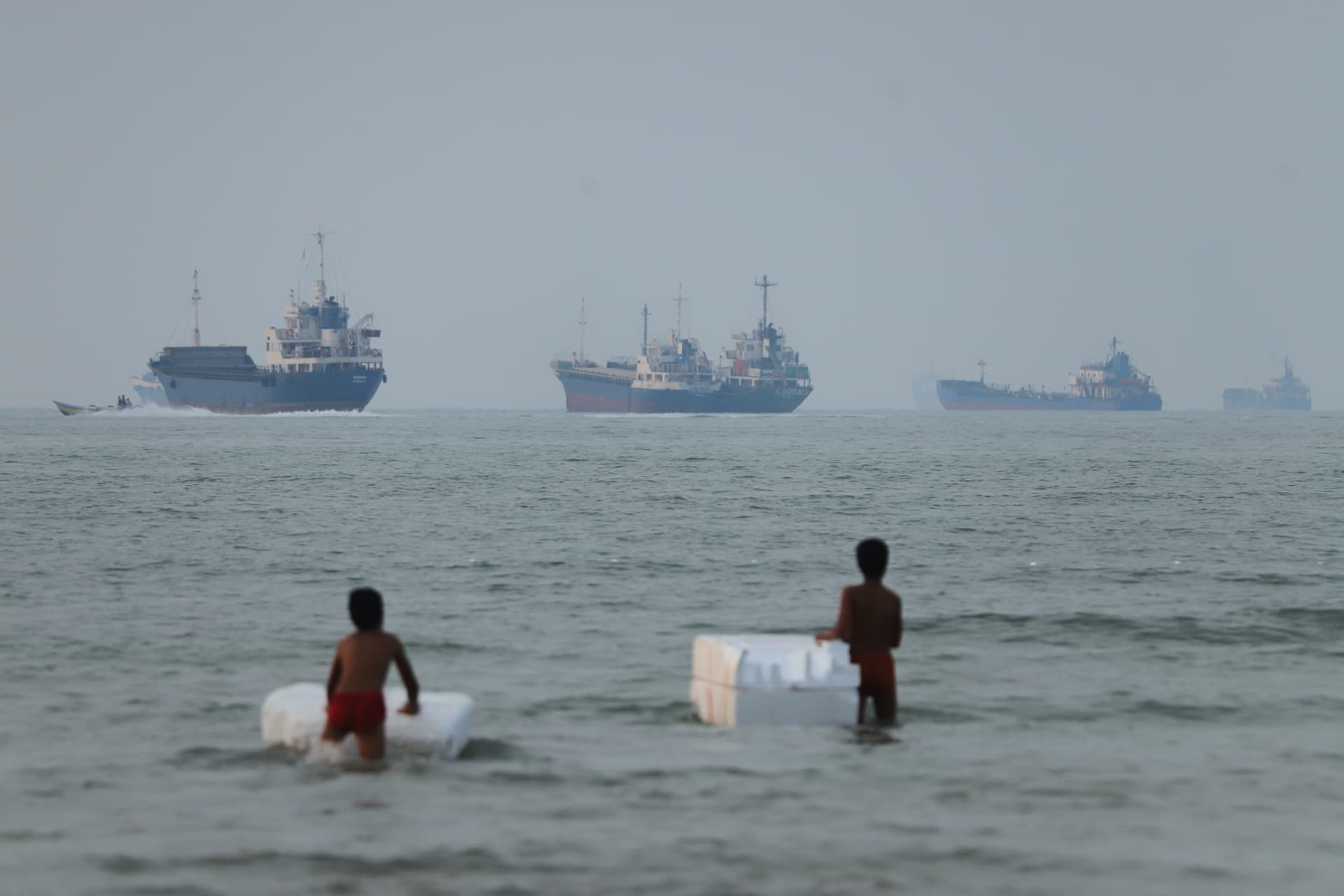 Two boys stand play with foam floats as cargo ships and other vessels are seen in the Strait of Hormuz off Bandar Abbas, Iran, on June 30, 2026.
