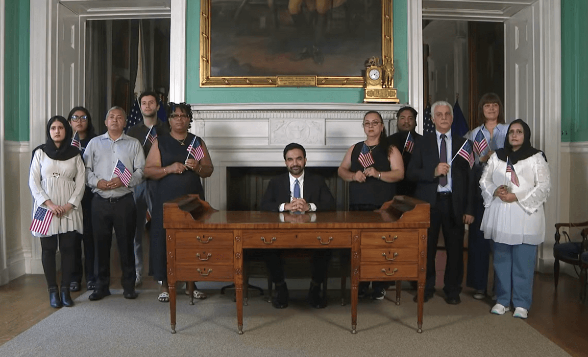 Mayor Zohran Mamdani, seated at George Washington's desk and surrounded by new citizens, delivers his Independence Day Eve speech.