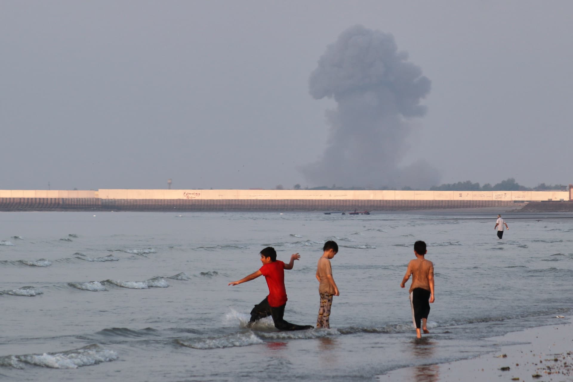 Razieh Poudat/ISNA via AP Three boys play in the shallow waters of the Strait of Hormuz, as a plume of smoke rises from an explosion in the background, off Bandar Abbas, Iran, July 13, 2026.